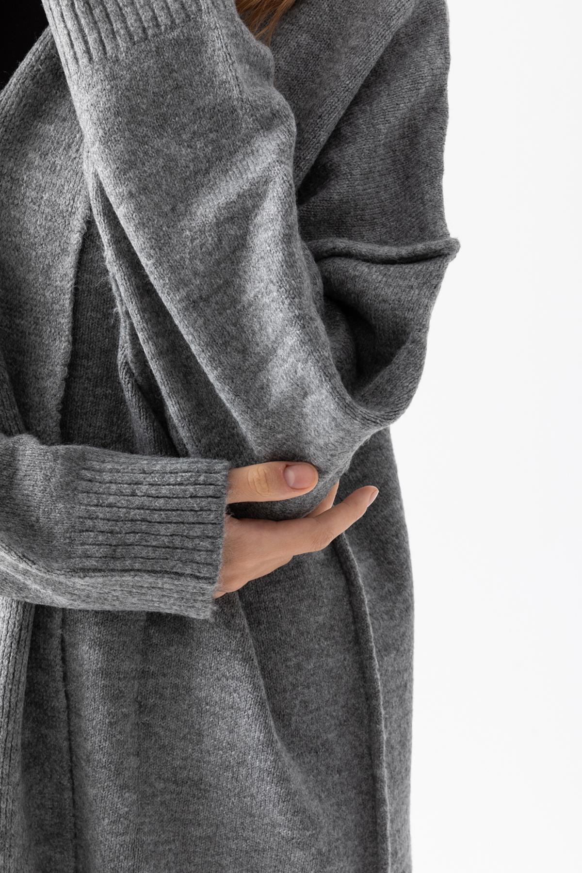 Könnte beinhalten: Langer, grauer Cardigan aus Strick. Der Pullover hat gerippte Bündchen und eine drapierte Front. Das Material wirkt weich und die Passform ist locker. Die Ärmel sind lang und das Design ist schlicht und elegant.